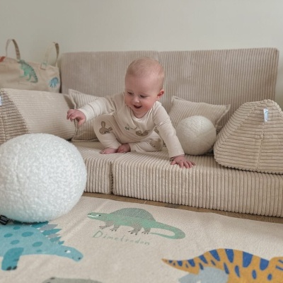 Bebé sentado em sofá infantil bege com almofadas triangulares e redondas, tapete com dinossauros no chão