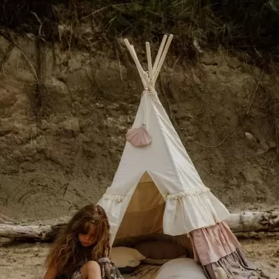 Tenda tipi branca com franjas cor de rosa e varas de madeira numa área de areia com menina sentada à frente