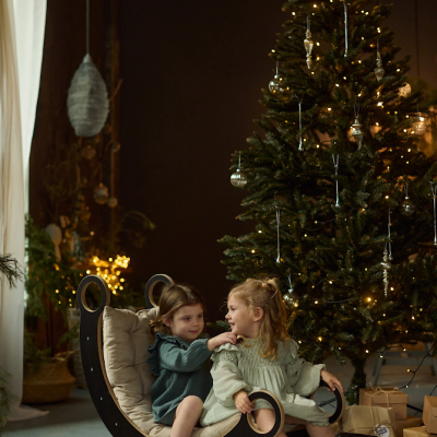 Duas crianças sentadas num baloiço de madeira perto de árvore de Natal com luzes e presentes.