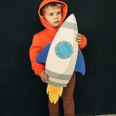 Criança com hoodie laranja segura brinquedo de foguetão de papelão com asas azuis.