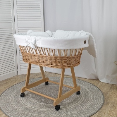 Berço de bebé em vime com base de madeira e rodas, forrado em branco, sobre tapete cinza circular com planta ao lado