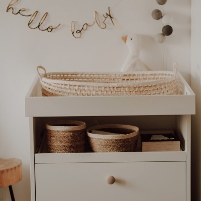 Móvel branco com gavetas, cestos castanhos, berço de vime, decoração de parede com texto 'hello baby' e pendentes de bolas cinzentas