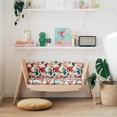Sofá de madeira clara com almofadas florais suspenso, estante branca com decoração e planta em vaso ao lado