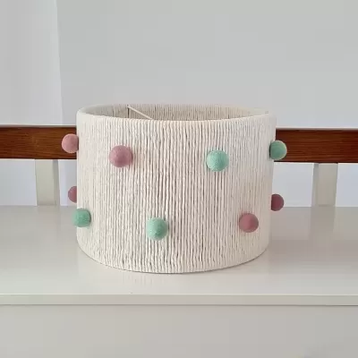 Cesto branco com pompons rosa e verdes sobre cómoda branca com puxadores de madeira.