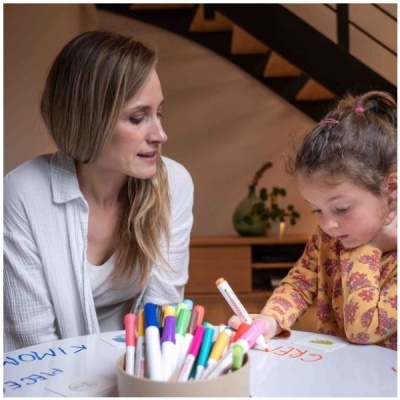 Menina a pintar com marcador cor-de-rosa à mesa com vários marcadores junto a mulher em ambiente interior