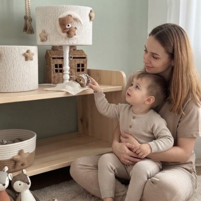 Candeeiro de mesa branco com ursinho e estrelas decorativas numa prateleira de madeira, mãe e filho sentados no chão a brincar