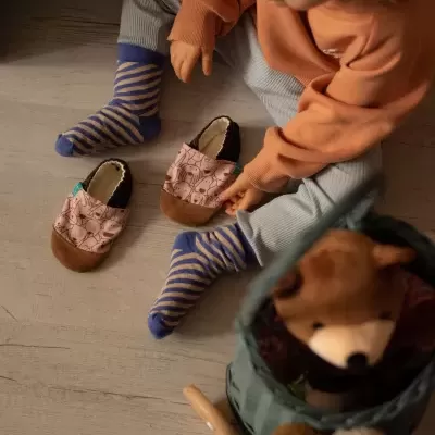 Criança com meias riscas azul e castanho e pantufas cor-de-rosa com padrão, junto a um cesto verde com peluche