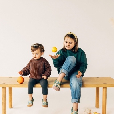 Duas crianças sentadas num banco com sapatos verdes com padrão de fruta e roupas casuais.