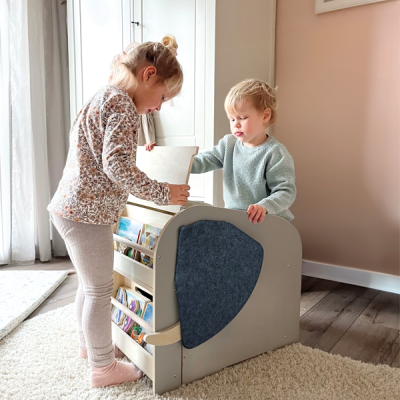Estante infantil de madeira cinzenta com livros e feltro azul num quarto com tapete creme