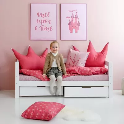 Cama branca com almofadas e decoração rosa, parede rosa com quadros, criança sentada na cama.
