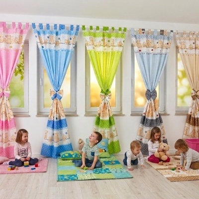 Cinco cortinas infantis coloridas com estampas de ursos penduradas em janelas numa sala com crianças sentadas em tapetes coloridos.