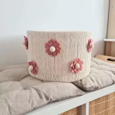 Cesto decorativo redondo com flores cor-de-rosa em tecido bege, sobre almofadas em prateleira