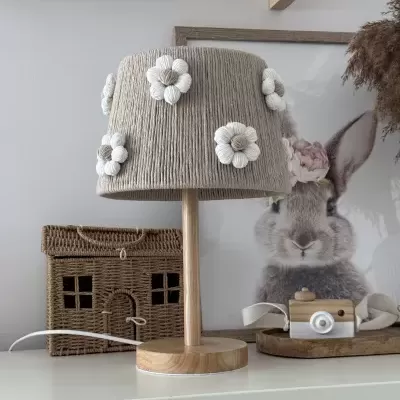 Abajur de madeira clara com cúpula bege decorada com flores brancas ao lado de caixa de vime e brinquedo de coelho