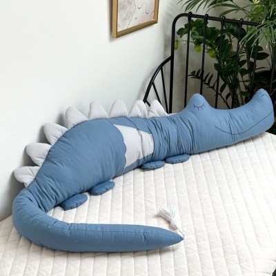 Almofada em forma de crocodilo azul em cama branca com moldura preta e plantas ao fundo
