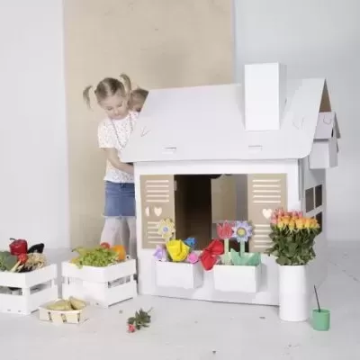 Casa de brincar branca de cartão com flores e menina ao lado
