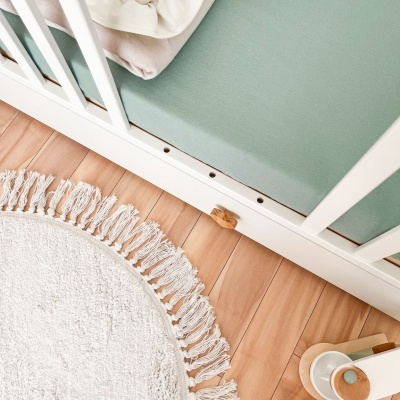 Berço branco com colchão verde menta, tapete bege com franjas em chão de madeira clara.