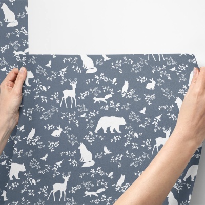 Papel de parede azul com padrão branco de animais da floresta e folhas, mãos a segurá-lo.