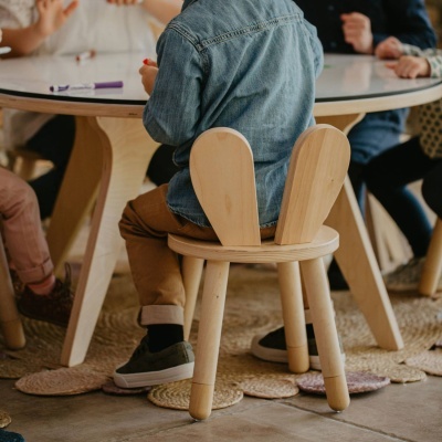 Criança sentada numa cadeira de madeira com encosto em forma de orelhas de coelho junto a mesa redonda