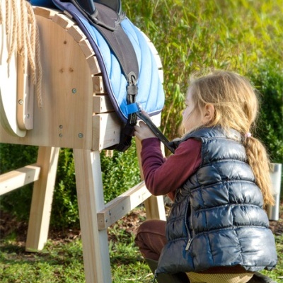 Criança com colete azul escuro junto a sela azul clara num cavalo de madeira ao ar livre
