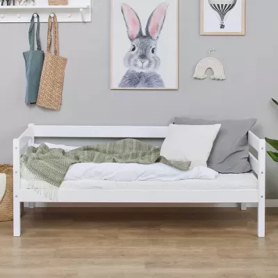 Cama de criança branca com mantas e almofadas num quarto decorado com quadros e sacos pendurados