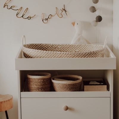 Cômoda branca com almofada trançada, cestos trançados, brinquedo cisne branco e decoração 'hello baby'