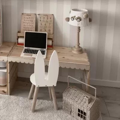 Secretária de madeira clara com cadeirão infantil branco, computador portátil, candeeiro de mesa com coruja e cesta tecida em forma de casa