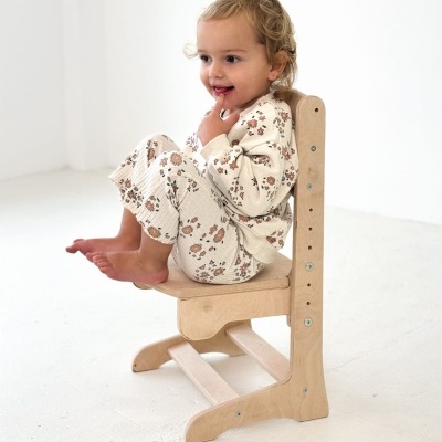 Criança com pijama floral sentada numa cadeira de madeira clara ajustável em ambiente branco