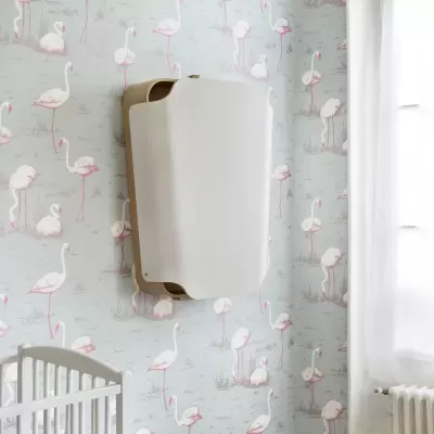 Móvel suspenso branco com base de madeira clara numa parede com papel de parede de flamingos rosa ao lado de um berço branco e janela com cortina branca