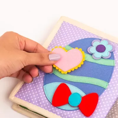 Livro infantil tátil com peças de tecido colorido, capa lilás às bolinhas e coração de velcro sendo segurado por mão