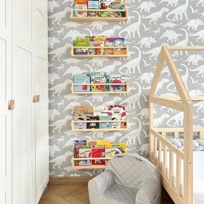 Quarto infantil com papel de parede de dinossauros, estantes de madeira com livros, cadeira pequena cinza e cama de madeira clara.