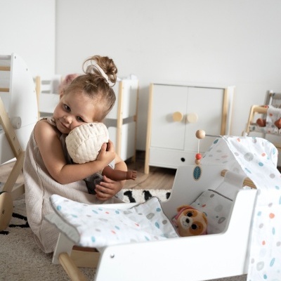 Menina com boneca junto a berço branco de madeira com tecido colorido num quarto claro