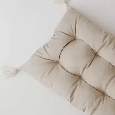 Almofada retangular bege com tassels, sobre superfície branca e fundo de azulejos brancos.