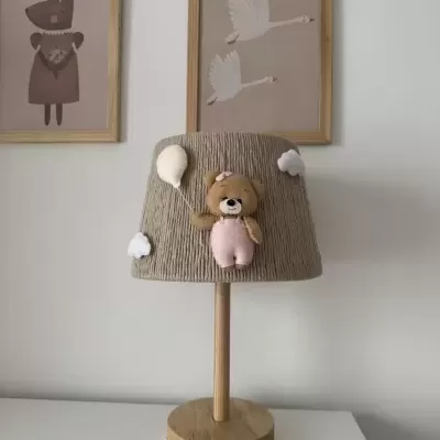 Candeeiro de mesa com abajur cinzento decorado com ursinho e balão, base em madeira, em mesa branca, com quadros decorativos na parede branca atrás.