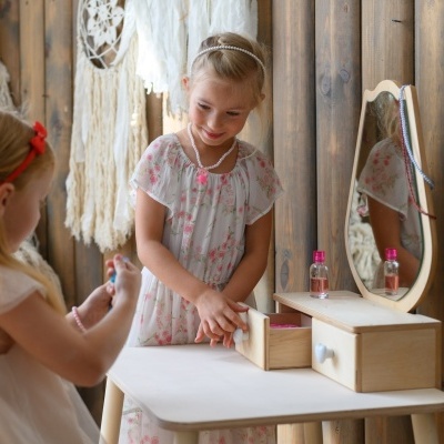 Duas meninas brincam com um tocador de madeira clara com espelho oval numa sala com paredes de madeira escura.