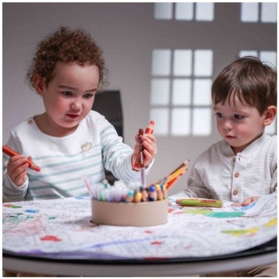 Duas crianças pequenas a pintar uma grande folha de desenho numa mesa redonda com lápis de cor e canetas.