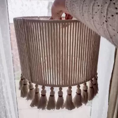 Abajur de tecido bege com pompons pendurados e mão segurando-o