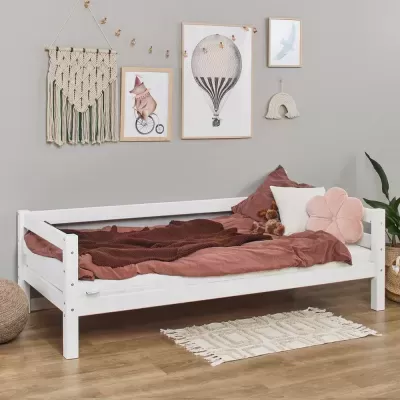 Cama de criança branca com roupa de cama castanha e almofadas rosa e branca num quarto com decoração em macramé e quadros na parede