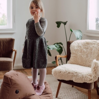 Menina com vestido cinzento e pantufas cor-de-rosa numa divisão clara com poltrona e plantas