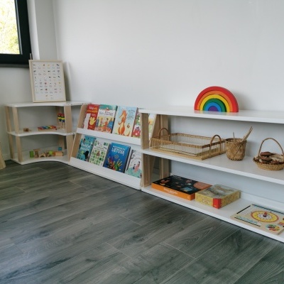 Estante branca com livros infantis, brinquedos e cestos, em ambiente luminoso com chão escuro e paredes brancas.