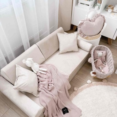 Sofá infantil bege com almofadas e manta rosa, cadeira de vime com boneca, tapete redondo branco e rosa com pompons em quarto infantil.