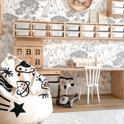 Quarto infantil com prateleiras de madeira, caixas transparentes, papel de parede com animais, puff com estampas pretas, cadeira branca, tapete branco, mini bicicleta e casinha de madeira.