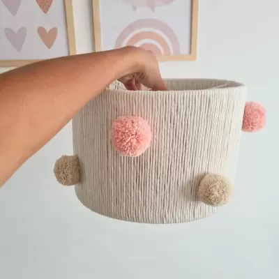 Cesto redondo em tecido bege com pompons rosa e bege segurado à frente de parede com quadros
