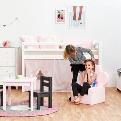 Quarto infantil com cama branca elevada, mesa de brincar rosa com cadeiras pretas, móveis brancos e tapete cor de rosa.
