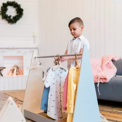 Criança com camisa branca a mexer numa estrutura de madeira com roupas penduradas numa sala