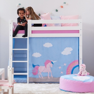 Cama alta branca para crianças com decoração de unicórnio e arco-íris, junto com puff e peluche cor-de-rosa.