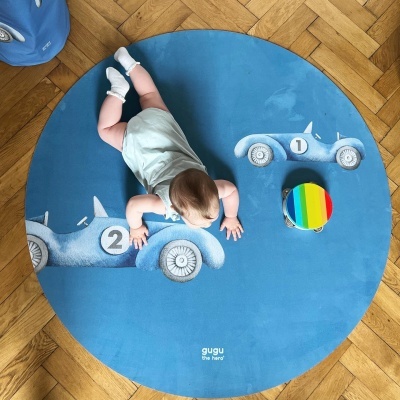 Tapete redondo azul com carros de corrida cinzentos e bebé deitado com tambor colorido perto