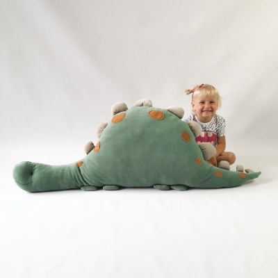 Peluche grande em forma de dinossauro verde com manchas castanhas e espigões cor de areia, com criança ao lado