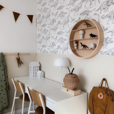 Quarto infantil com secretária, cadeiras, candeeiro, prateleira redonda com dinossauros e papel de parede com padrão de dinossauros.