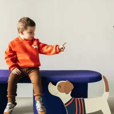 Criança de hoodie laranja e calças castanhas sentada num banco azul, apontando para brinquedo de cão em madeira.