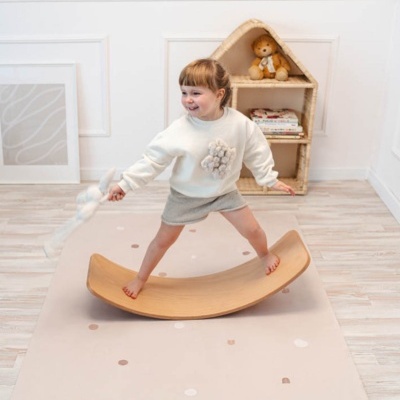 Criança em prancha de equilíbrio de madeira sobre tapete bege com pontos neutros, prateleira em forma de casinha ao fundo.